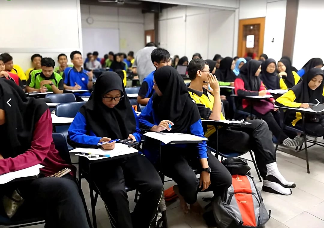 A classroom full of students in yellow and blue uniforms sitting at desks during a lecture or exam, with diverse groups scattered throughout the room